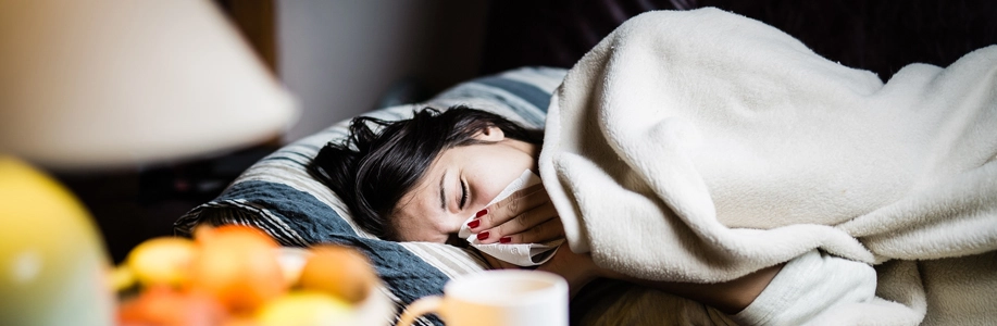 Sick woman wrapped in blanket on couch blowing nose